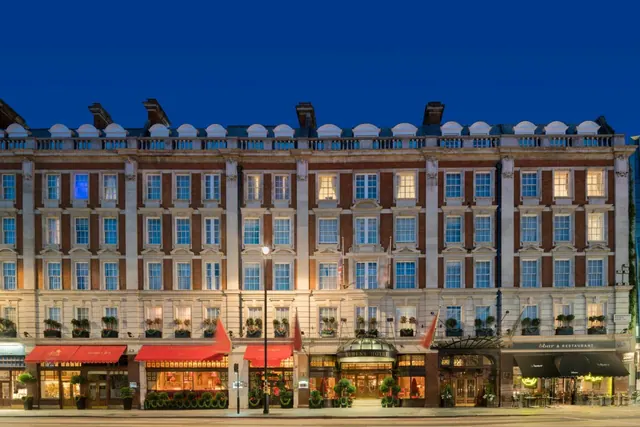 Evening exterior view of a multi-storey brick and stone hotel building with lit windows, red awnings and a restaurant at street level.