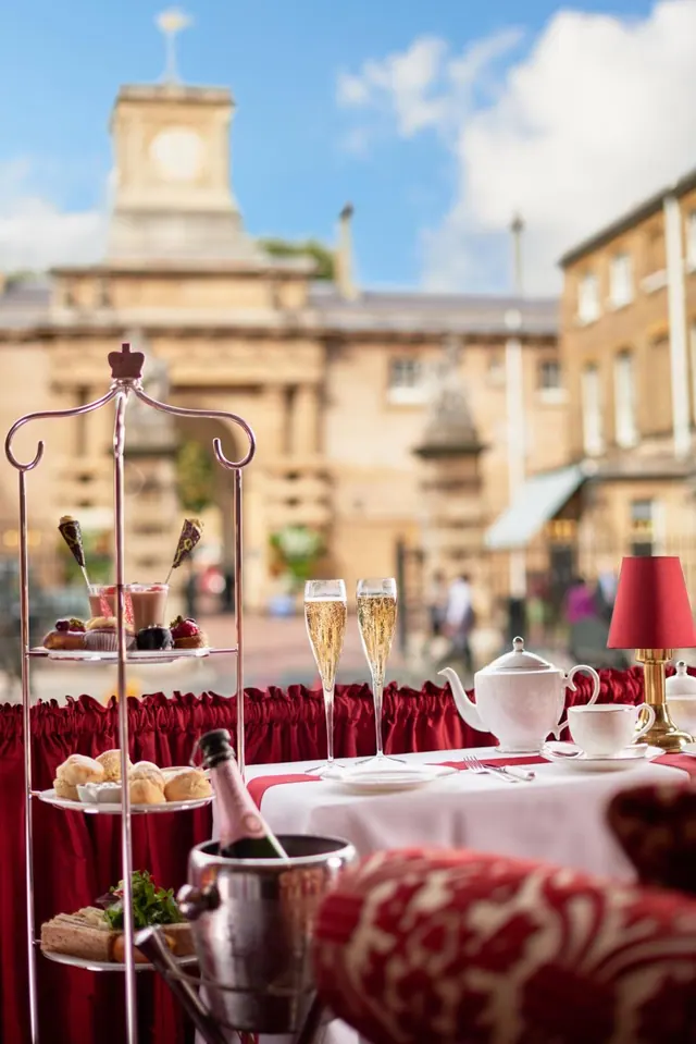 Afternoon tea is set out on an outdoor terrace table with champagne flutes, a teapot, tiered cake stands and a champagne bucket, with a historic stone building and clock tower behind.