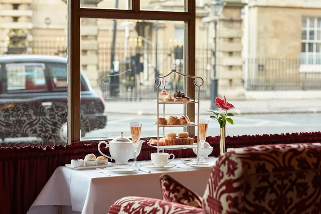 A table set for afternoon tea by a window, with a tiered stand of cakes and pastries, a teapot, champagne flutes and an armchair, overlooking a street with a black taxi.