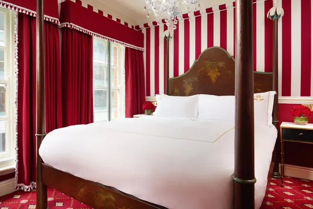 A bedroom with a four-poster double bed, red and white striped walls, red curtains, a chandelier and a red patterned carpet.
