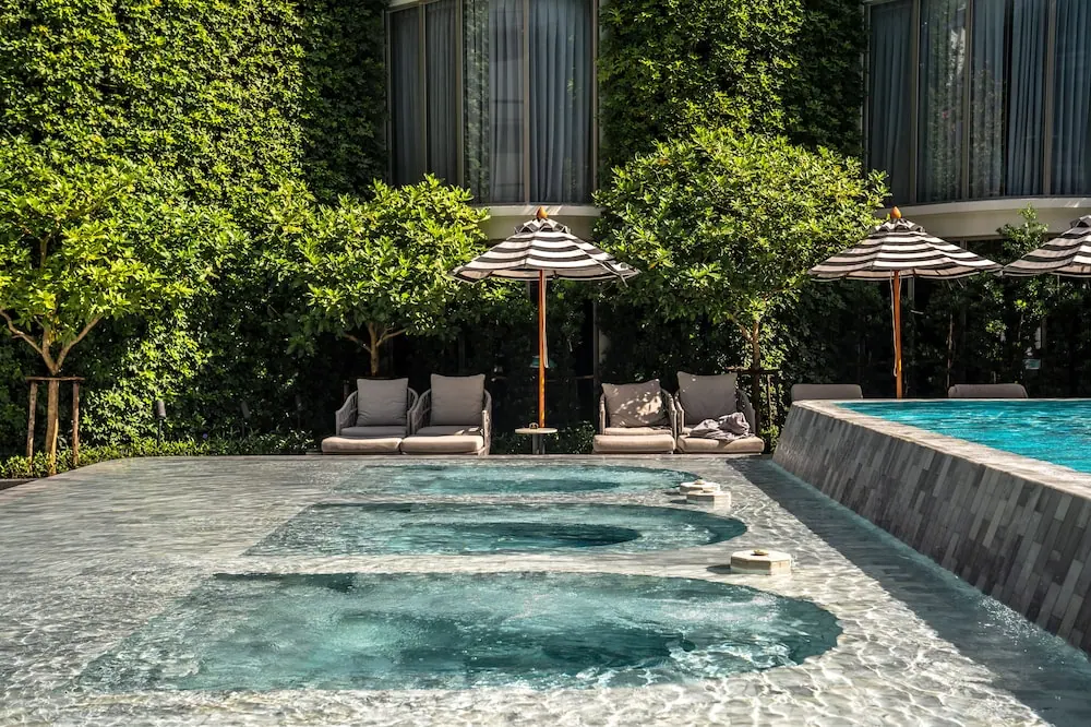 Three tiered shallow spa pools sit in front of a larger swimming pool, with striped umbrellas and grey loungers beneath green trees beside a building with curtained windows.