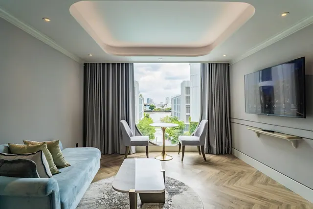 A living room with a light blue sofa, a round marble-top coffee table, a round side table with two grey chairs, a wall-mounted television, and a window with a view of a river and buildings.