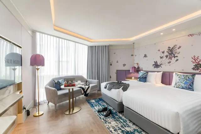 A hotel bedroom with two beds dressed in white linen, a grey sofa and round table by a sheer-curtained window, and a wall-mounted television.