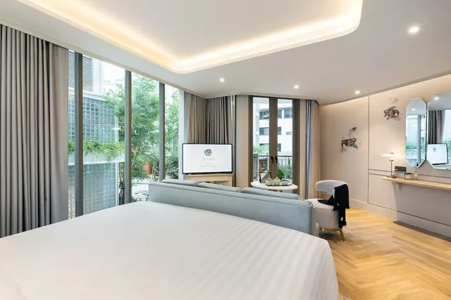 A bedroom with a large white bed, a grey sofa, a television on a stand and floor-to-ceiling windows looking onto trees and buildings.