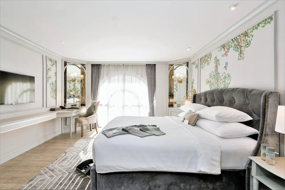 A bedroom with a made king bed under a grey tufted headboard, a wall-mounted television, a white desk with a chair and mirror, and an arched window with sheer curtains.
