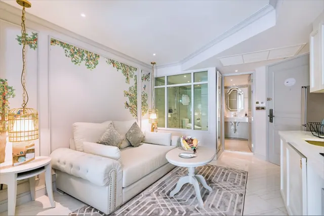 A white tufted sofa, a round side table and a kitchenette sit in a room with a painted tree mural, and the bathroom is visible through a doorway on the right.