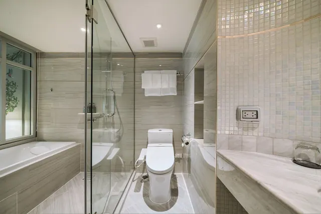 A bathroom with a bathtub beside a glass-walled walk-in shower, a toilet, white towels on a wall rail, and a marble vanity counter, with greenery visible through the window.