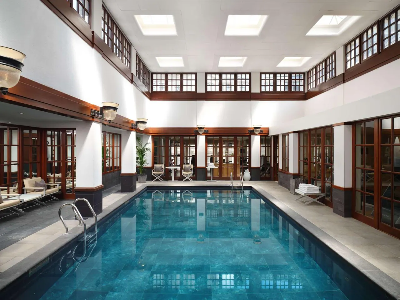 Indoor swimming pool under a row of skylights, with sun loungers, wooden-framed windows and tiled walls around it.