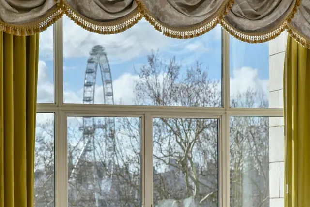 A window framed by gold curtains and a pelmet looks out past bare trees to the London Eye under a partly cloudy sky.