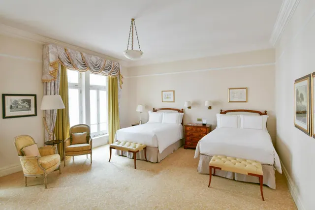 A bedroom with two beds dressed in white linen, two armchairs by tall windows hung with patterned drapes, and a patterned carpet.