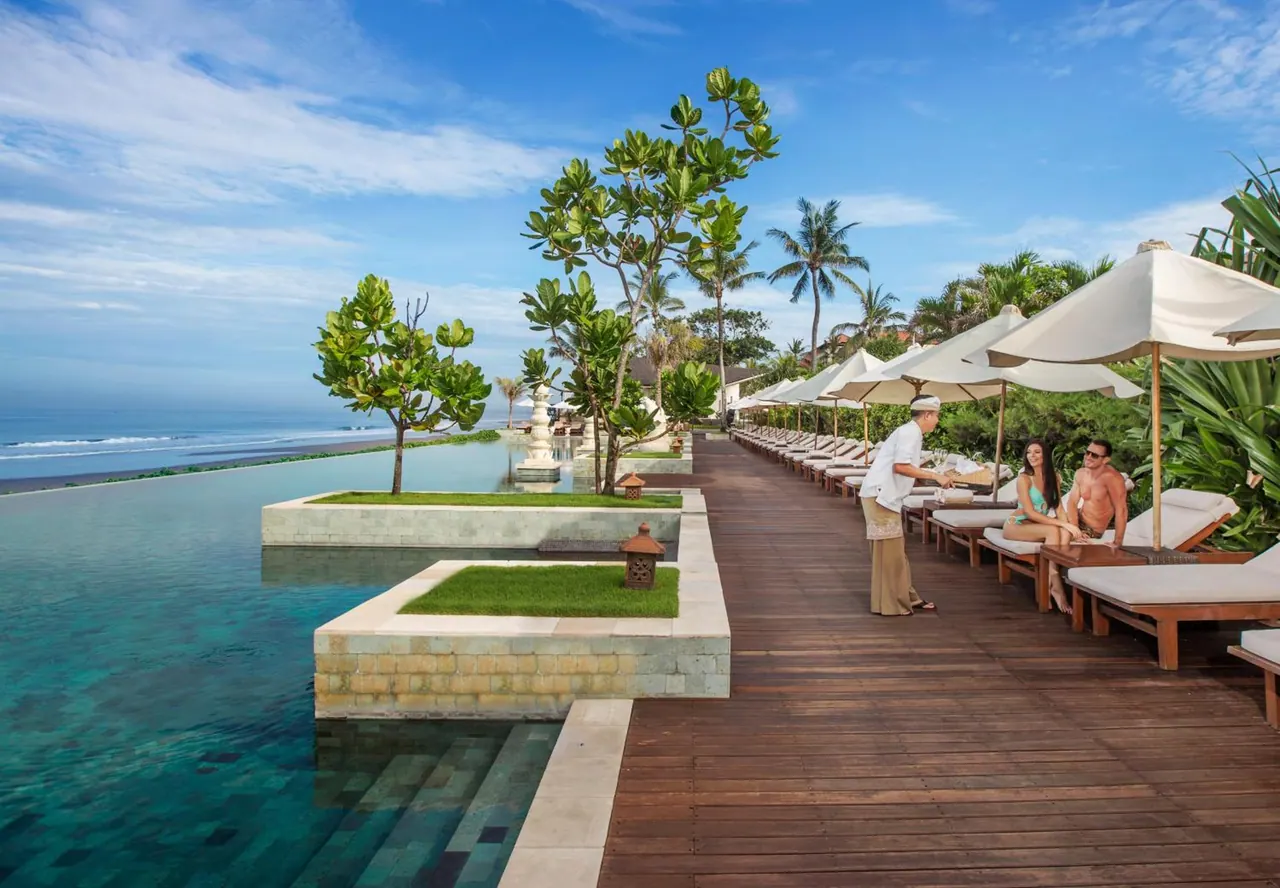 An infinity-edge pool runs alongside a wooden deck lined with sun loungers and parasols, with the sea and beach visible beyond, while a staff member attends two seated guests.