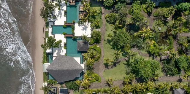 Aerial view of a beachfront resort with long turquoise pools, thatched pavilion roofs, landscaped gardens and the ocean along the left edge.