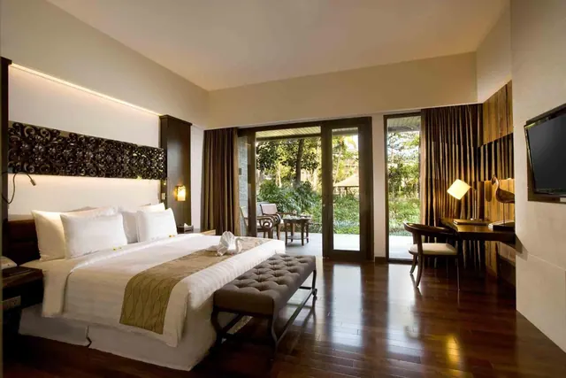 A bedroom with a king bed, a tufted bench at the foot, a wooden desk and wall-mounted television, with glass doors opening onto a garden terrace.