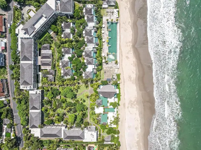 Aerial view of a beachfront resort with rooftops, tree-lined gardens, a long swimming pool and a sandy beach beside the surf.