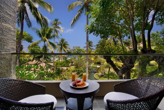 A balcony with two wicker chairs and a round table holding a plate of fruit and two glasses of juice, overlooking palm trees and a garden with a pond.