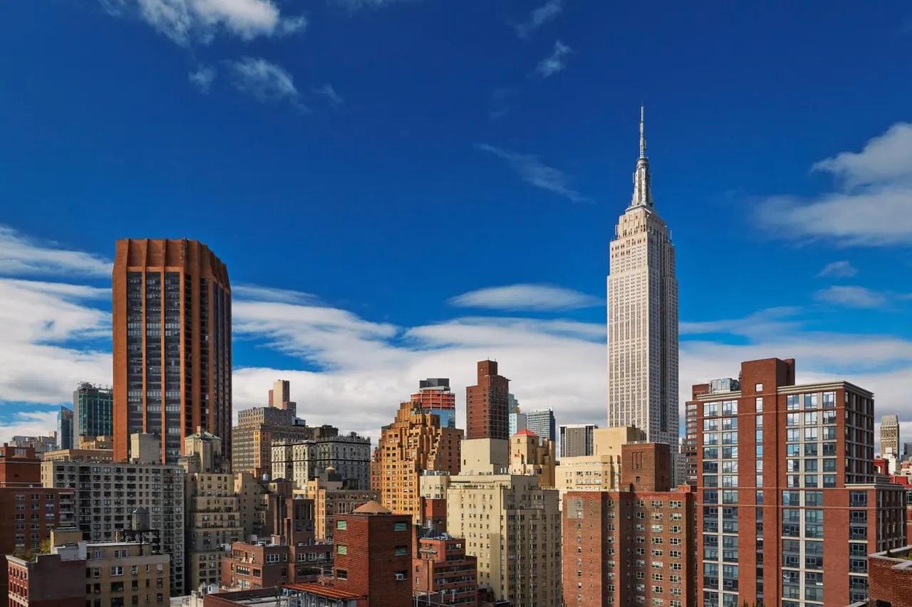 A view over Midtown Manhattan rooftops toward the Empire State Building under a blue sky with scattered clouds.