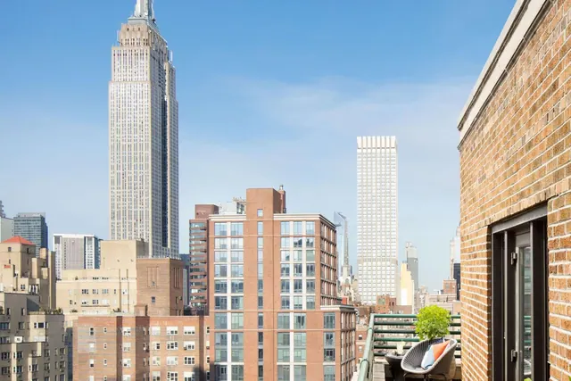 A brick-walled terrace with a metal railing, a chair with a cushion and a potted plant, looking out over city buildings including the Empire State Building.