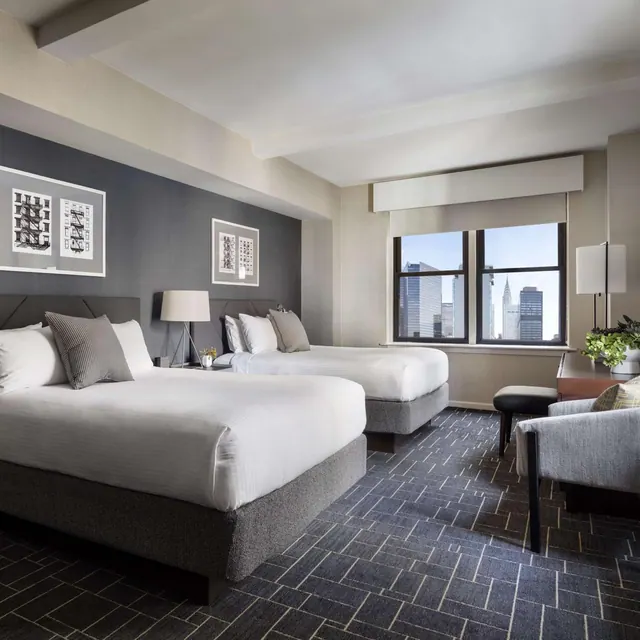A bedroom with two made beds, a desk and an armchair, with a window showing city skyscrapers including the Chrysler Building.