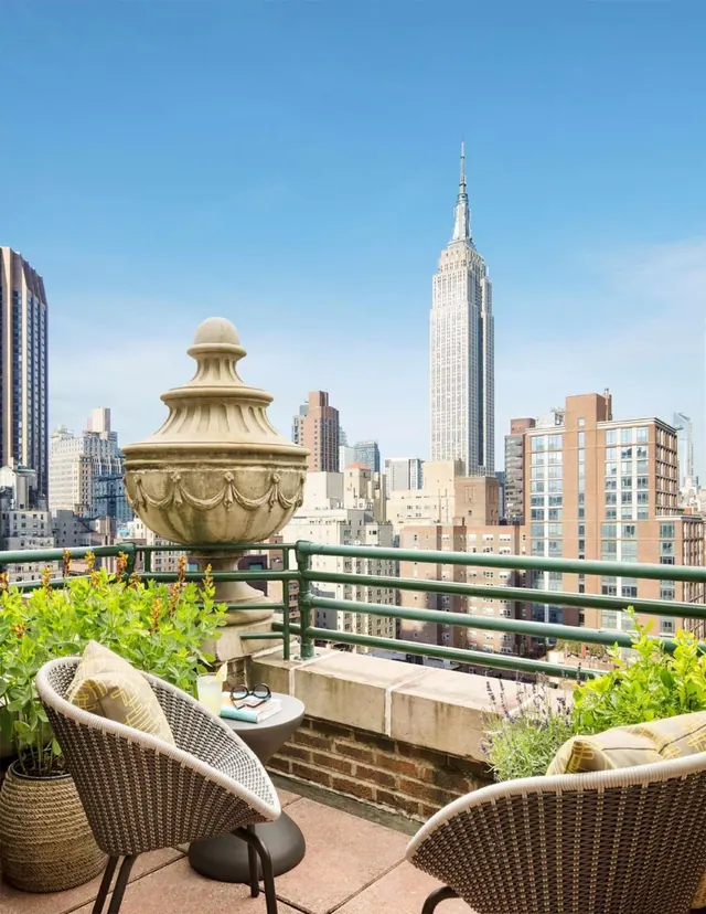 A rooftop terrace with wicker chairs, a stone urn and potted plants overlooks city buildings and the Empire State Building under a clear blue sky.
