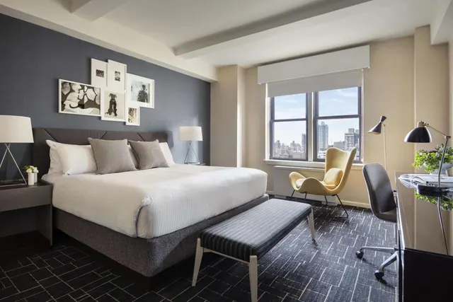 A staged hotel bedroom with a king bed, a yellow armchair by the window, and a desk with a lamp, with city buildings visible outside.