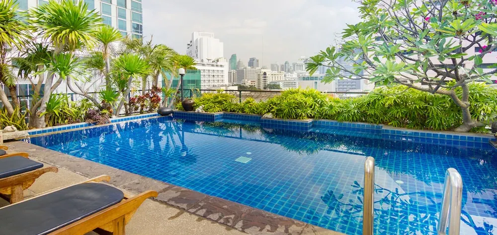 A rooftop swimming pool with blue tiles, sun loungers at the left edge, tropical plants, and a city skyline in the background.