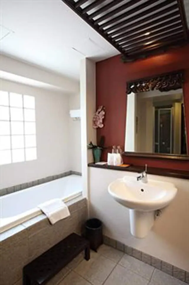 A bathroom with a built-in bathtub beside a grid window, a pedestal sink under an ornate mirror, and a red accent wall.