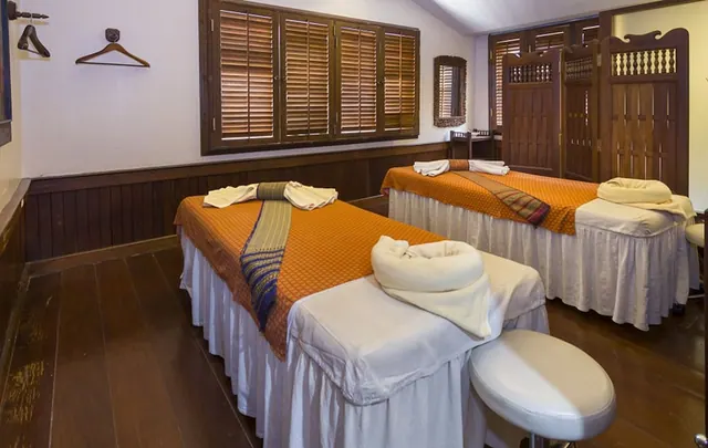 Two massage beds with orange covers and folded white towels sit in a wood-panelled treatment room with shuttered windows.