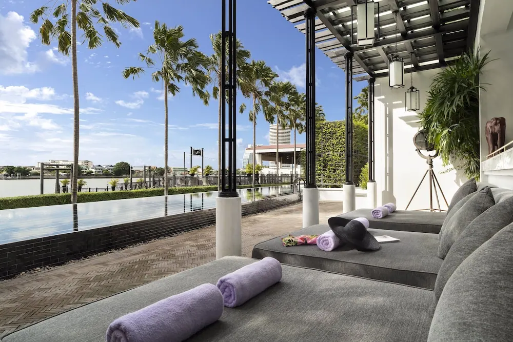 Rolled lavender towels lie on grey sun loungers beside an infinity pool, with a river, palm trees and buildings beyond.