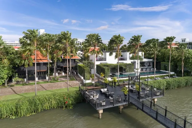 An elevated view of a riverside hotel deck with outdoor lounge and dining furniture, a swimming pool with loungers, and palm trees beside a river.