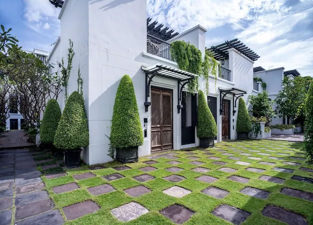 White villa facades with black pergolas, cone-shaped topiary and a stepping-stone path set in grass.
