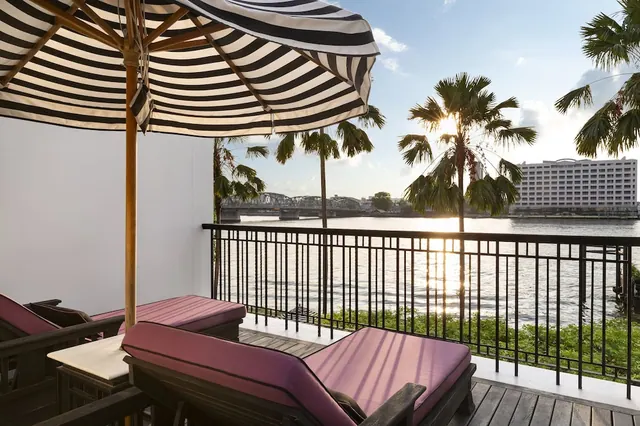 A private balcony with two purple-cushioned loungers and a striped umbrella overlooks a river with palm trees and a bridge at sunset.