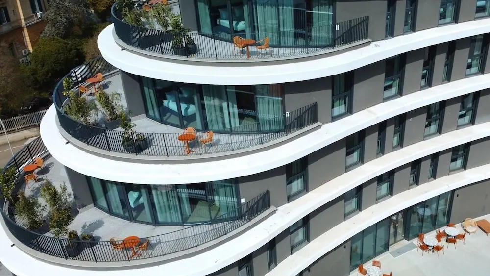 Aerial view of a curved multi-storey building facade with balconies furnished with orange tables and chairs, and a ground-level terrace with outdoor seating.