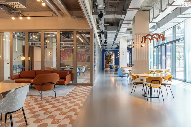 A modern open lounge with orange sofas and armchairs, yellow dining chairs at round tables, and glass walls and tall windows along one side.