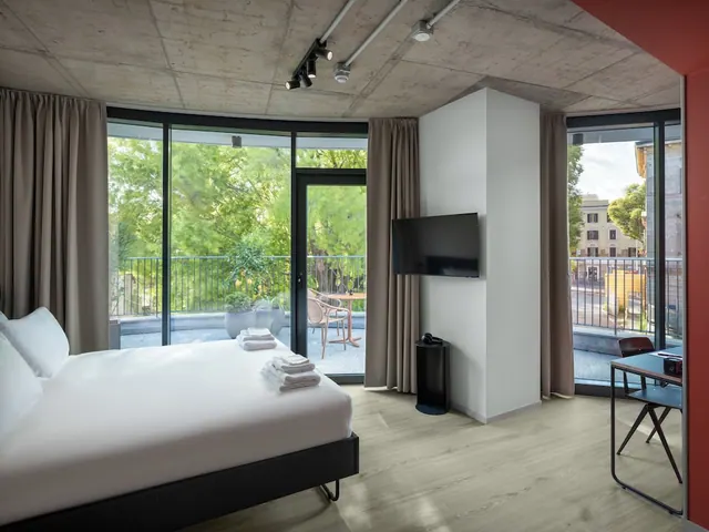 A bedroom with a made king bed, a wall-mounted television, and floor-to-ceiling glass doors opening onto a balcony with chairs and a table, with trees beyond.
