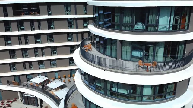 Aerial view of a curved modern building with balconies furnished with orange chairs and tables, and a terrace below with white umbrellas over outdoor seating.