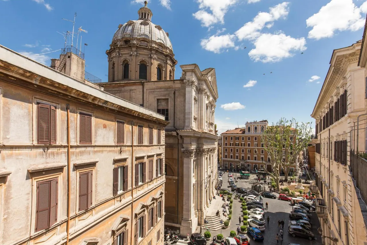 View from an upper window over a city square with a domed church, parked cars along the street, and multi-storey buildings under a partly cloudy blue sky.