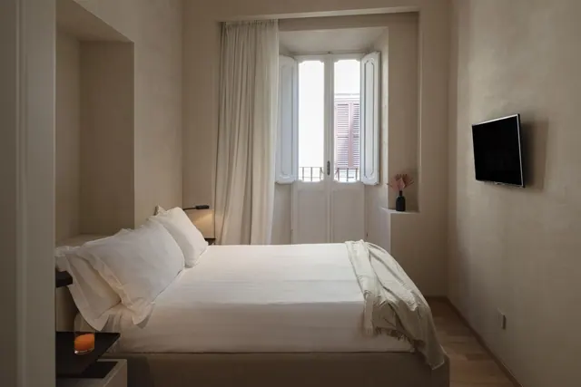 A bedroom with a made double bed, a wall-mounted television, and a window with open shutters facing a neighbouring building.