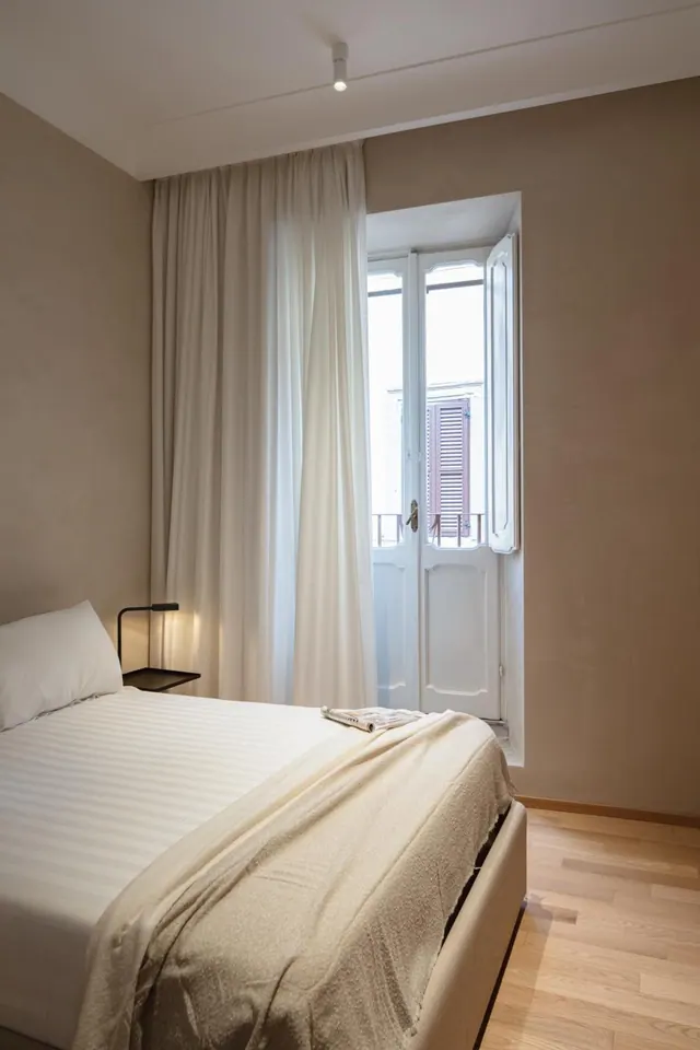 A bedroom with a made double bed, sheer cream curtains, and open French doors onto a small balcony with a railing.