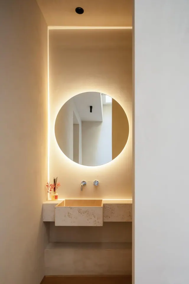 A bathroom with a round backlit mirror above a stone basin on a wall-mounted vanity, with a wall-mounted tap and a reed diffuser on the ledge.