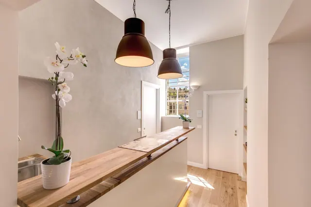 A wooden kitchen counter with a sink, two dark pendant lamps and a white orchid, with a tall window showing buildings beyond.