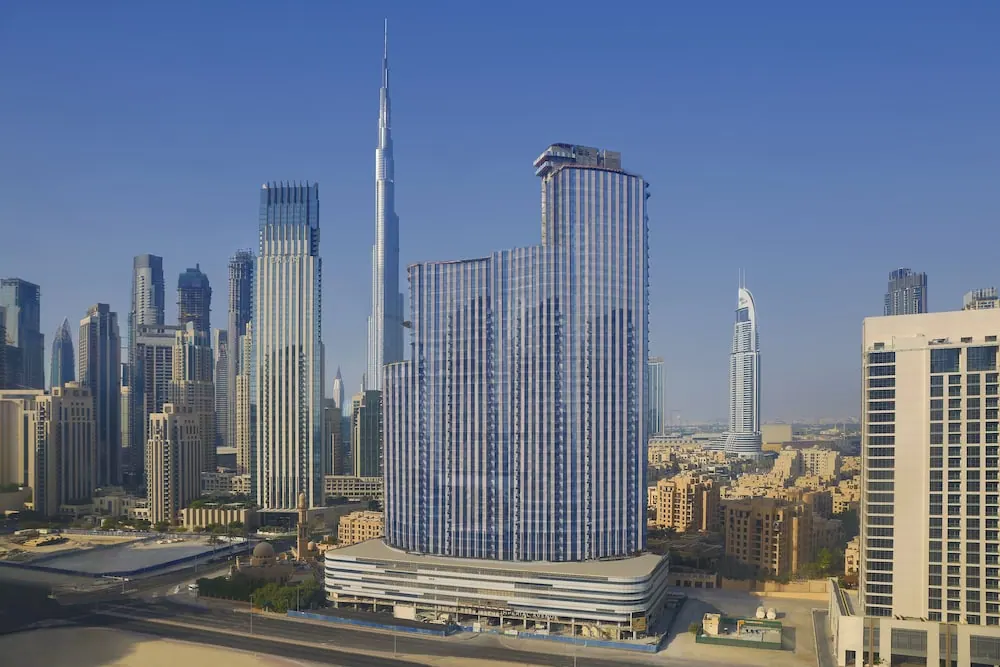 Elevated view of a tall glass hotel tower in Dubai with the Burj Khalifa and other skyscrapers rising behind it under a clear blue sky.