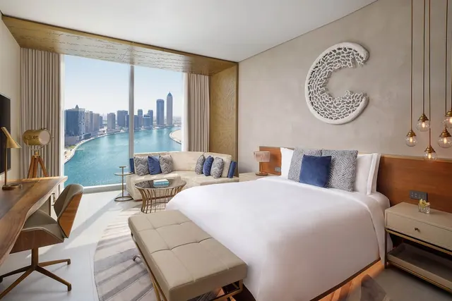 A bedroom with a large white bed, a sofa and a work desk, and floor-to-ceiling windows looking over a canal and a city skyline.