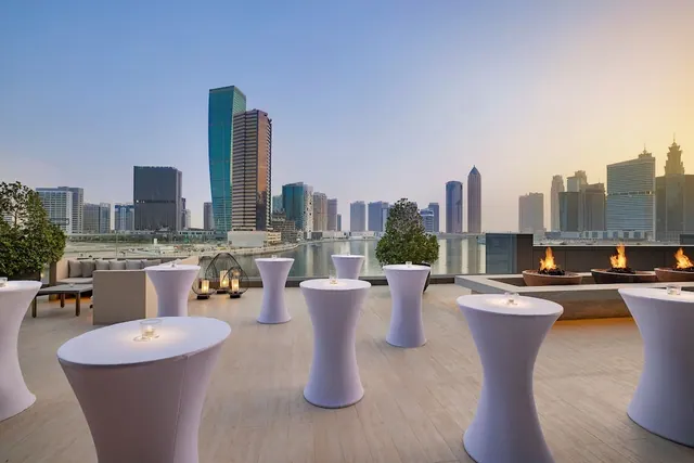 A rooftop terrace with white cocktail tables, lit candles, gas fire pits and a view over a canal and city skyline towers at dusk.