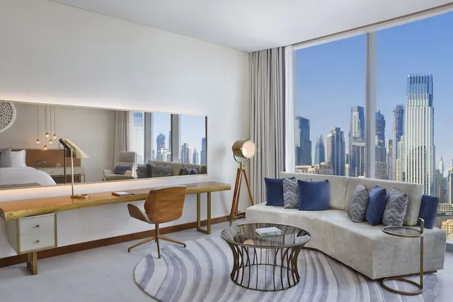 A living area with a curved sofa, a wooden desk and chair, and a floor-to-ceiling window looking onto a city skyline of high-rise towers, with a bed visible in the mirror.