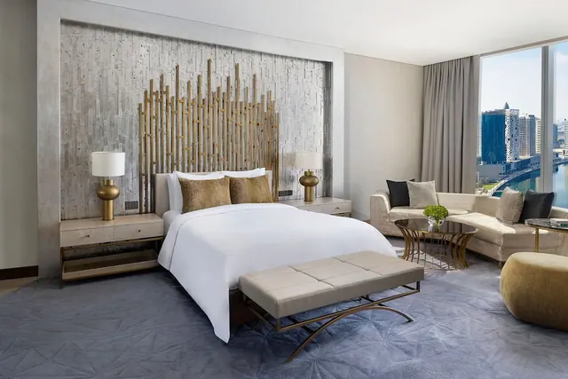A bedroom with a king bed, a gold slatted headboard, a seating area and floor-to-ceiling windows looking out over city towers and water.
