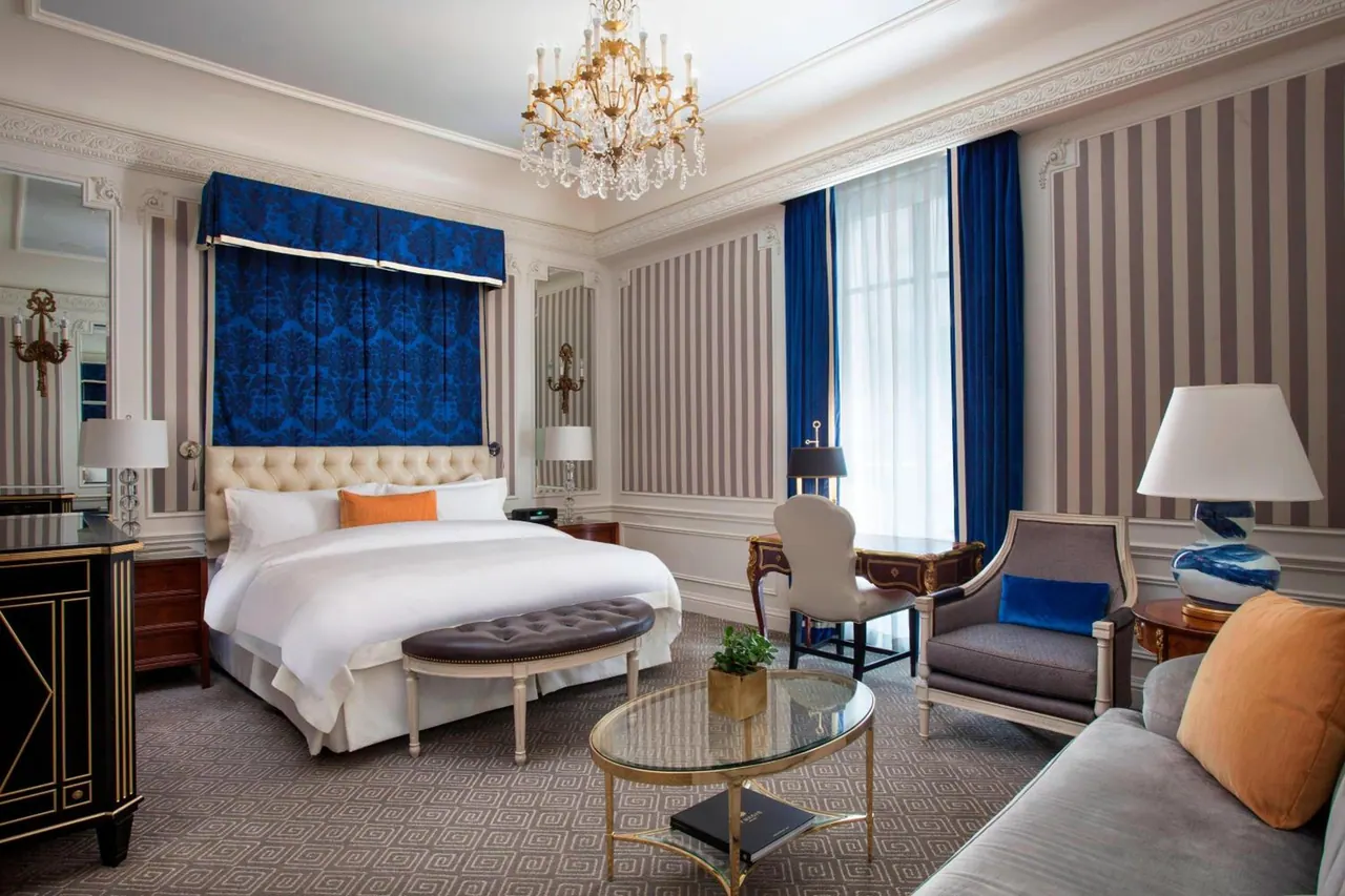A large bedroom with a made king bed under a blue damask headboard, a crystal chandelier, striped walls, an armchair and sofa seating area, and a small desk by a tall window with sheer curtains.