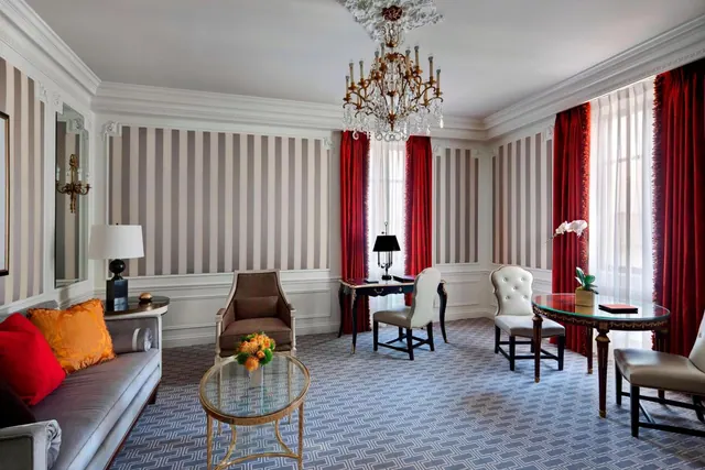 A furnished hotel suite living area with striped walls, a crystal chandelier, red curtains, a sofa, armchairs, a writing desk and a round table.