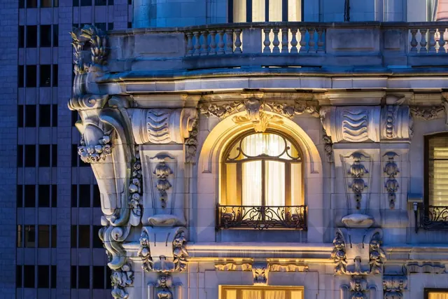 Ornate stone facade of a building at dusk with an illuminated arched window and a wrought iron balcony railing.