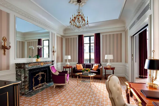 A formal living area with striped walls, a crystal chandelier, a marble fireplace, a sofa with orange cushions, and a writing desk in the foreground.