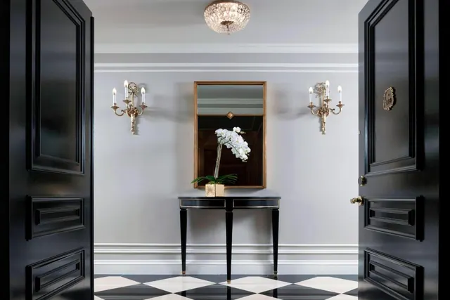 A symmetrical entrance foyer with open black double doors, a black and white checkered floor, a black console table holding a white orchid, a gilt mirror, wall sconces and a crystal ceiling light.